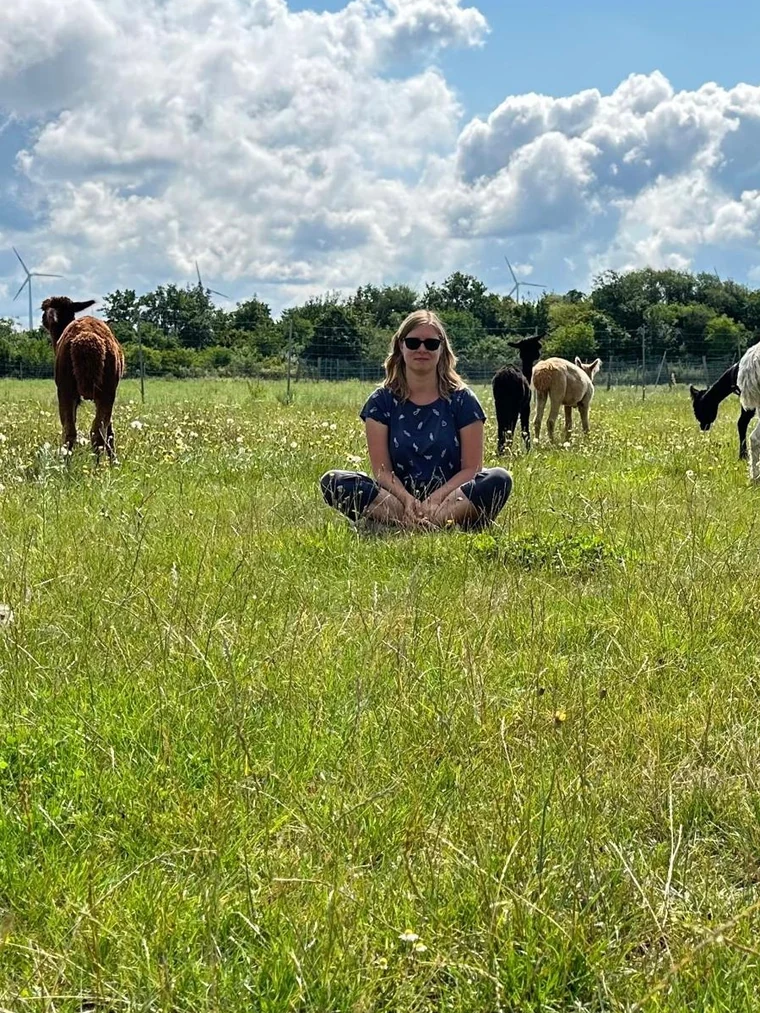 Jennifer Arbter sitzt in der Blumenwiese, ihre Alpakaherde grast rund um sie herum